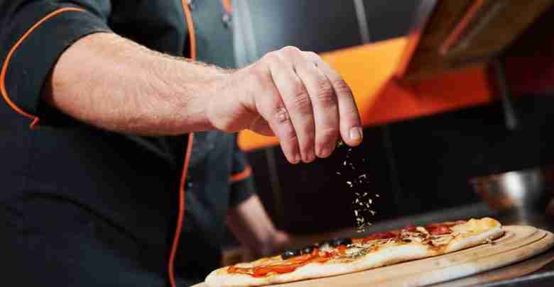 hand of chef baker in uniform adding spice into pizza after pizz
